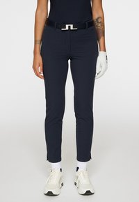 Persona che indossa pantaloni slim blu navy con spacchi laterali, cintura nera, guanto da golf bianco sulla mano destra, sneakers bianche e tatuaggi visibili sulle braccia.
