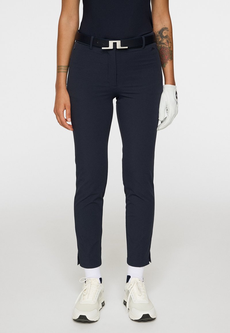 Persona che indossa pantaloni slim blu navy con spacchi laterali, cintura nera, guanto da golf bianco sulla mano destra, sneakers bianche e tatuaggi visibili sulle braccia.