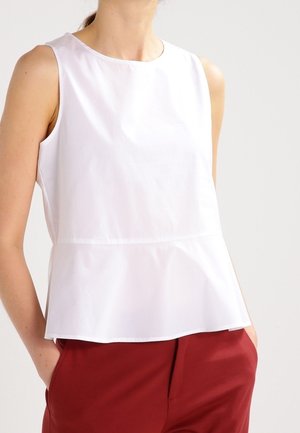 Femme portant un haut peplum blanc sans manches légèrement rentré dans un pantalon rouge taille haute, debout les mains dans les poches sur un fond clair.
