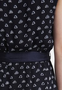 Robe noire à motifs de triangles irréguliers blancs, cintrée à la taille avec une ceinture bleu foncé, montrant un bras partiel sur le côté droit.