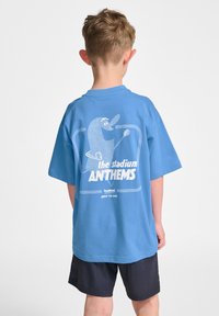 Blauwe katoenen T-shirt met een relaxed fit, featuring een witte grafiek van een personage en de tekst "de stadion ANTHEMS" op de achterkant.