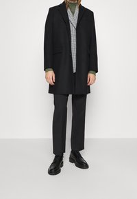 Sort frakke over en grøn turtleneck, parret med en ternet vest og sorte bukser. Har sorte snørebånds sko og en skræddersyet pasform.