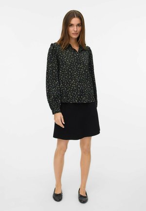 Kvinna stående i en svart blommig långärmad blus, svart kjol och svarta ballerinaskor mot en vit bakgrund.