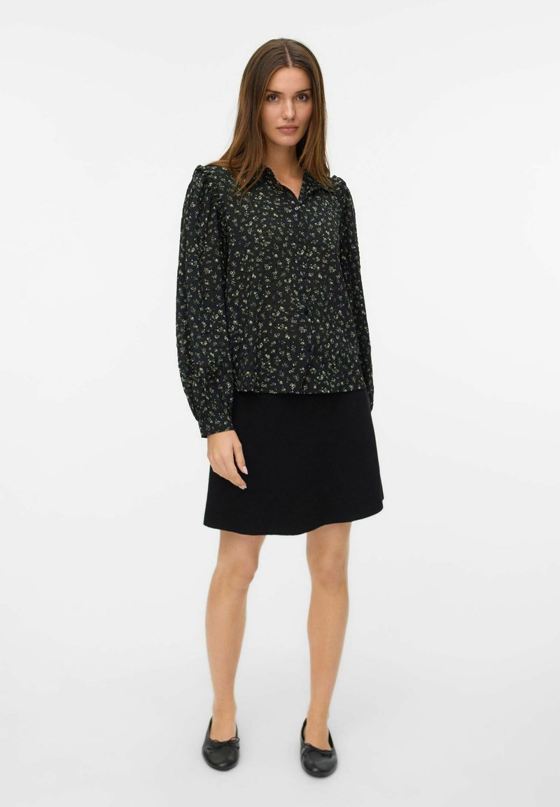 Donna in piedi con indosso una blusa nera a maniche lunghe con fantasia floreale, una gonna nera e ballerine nere su uno sfondo bianco.