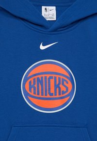 Modrá mikina s kapucí vyrobená z bavlněné směsi, se zpřehnanou grafiku velkého oranžového a modrého basketbalového míče s textem "KNICKS". Logo Nike zobrazené na hrudi.