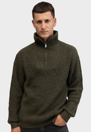 Homme portant un pull à fermeture éclair vert foncé et duveteux avec un col montant, debout avec une main dans la poche sur un fond blanc uni.
