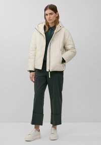 Off-white gesteppte Jacke mit Kapuze, ausgestattet mit einem Reißverschluss auf der Vorderseite und zwei Seitentaschen. Kombiniert mit dunkelgrünen Hosen und hellen Sneakers.