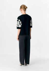 Femme avec les cheveux en chignon, portant un haut noir à manches courtes avec des broderies florales blanches, un jean sombre ample et des chaussures à talons blancs, se tenant de dos.