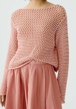 Personne portant un pull rose ample en maille ajourée à manches longues et une jupe plissée rose taille haute sur un fond uni.