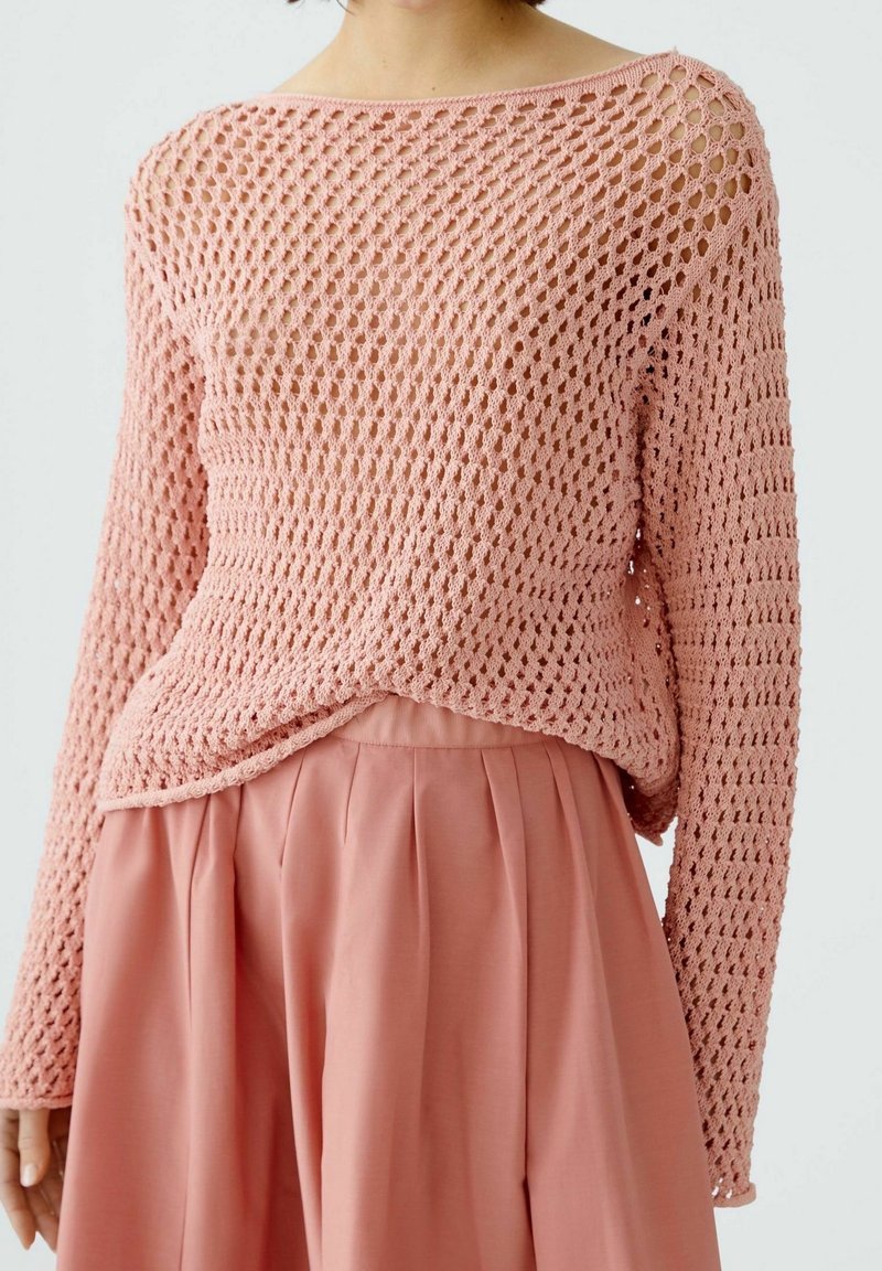 Personne portant un pull rose ample en maille ajourée à manches longues et une jupe plissée rose taille haute sur un fond uni.