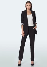 Kobieta stojąca pewnie w czarnym garniturze z wzorzystymi podwiniętymi rękawami, białą bluzką i czarnymi szpilkami na tle jednolitego tła.