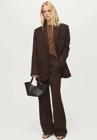 Brauner maßgeschneiderter Anzug mit weiten Hosen und einem übergroßen Blazer. Darunter ein geripptes braunes Oberteil. Hält eine kleine schwarze Handtasche.