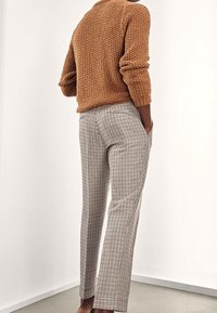 Personne portant un pull marron texturé et un pantalon à carreaux taille haute, debout les mains dans les poches devant un fond uni.