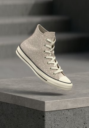 Lysebeige højtop sneaker med diskret leopardprint, hvide snørebånd og hvid sål med sort stribe, svævende over en betonhylde.