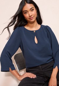 Donkerblauwe blouse met een ronde halslijn, een keyhole-opening en pofmouwen. Gladde textuur met een decoratief knoopdetail bij de hals.