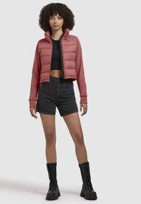 Rode cropped puffer jas met een hoge kraag, gevoerde voorkant en lange mouwen, gecombineerd met zwarte denim shorts en zwarte veterschoenen.