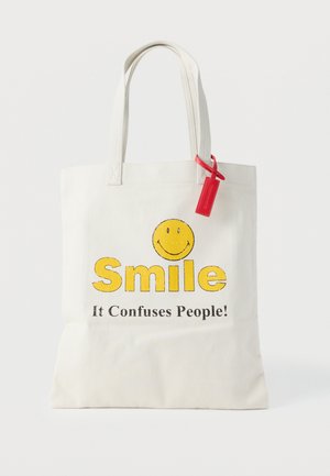 Sac fourre-tout blanc avec un visage souriant jaune et le texte « Souris, ça perturbe les gens ! », avec une étiquette rouge attachée à une des anses.