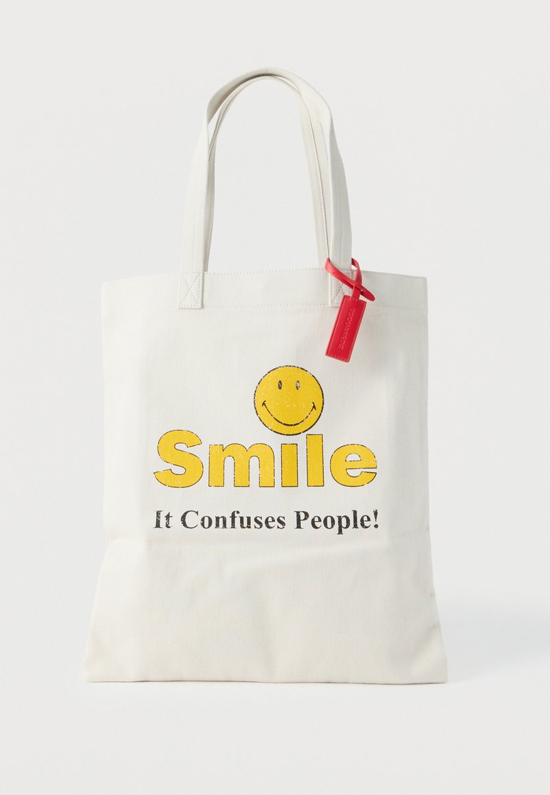 Λευκή τσάντα tote με κίτρινο χαμογελαστό πρόσωπο και το κείμενο "Χαμογέλα, μπερδεύει τους ανθρώπους!" και μια κόκκινη ετικέτα προσαρτημένη σε μια χειρολαβή.