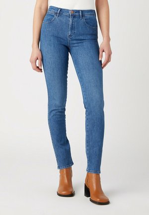 Kobieta w niebieskich dopasowanych jeansach i brązowych skórzanych botkach na obcasie na jasnym, jednolitym tle.