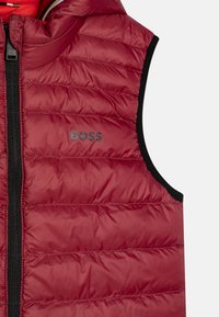 Gilet rembourré rouge avec fermeture éclair sur le devant, doté d'une bordure noire et du mot "BOSS" en gris sur la poitrine. Texture lisse et design matelassé.