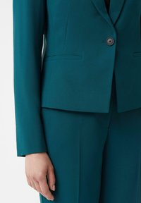 Teal tailored suit jacket with single black button and matching pants, worn on left side with hand relaxed by the side.