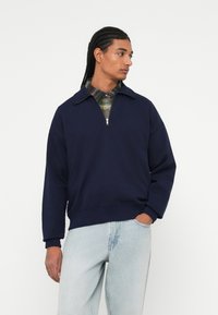 Navyblauwe sweatshirt met een ritskraag, voorzien van ribgebreide manchetten en zoom. Gelaagd over een groen-blauw geruit shirt. Lichte spijkerbroek.