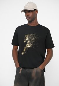 Man wearing beige cap and black t-shirt with gold pixelated horse graphic and text, paired with dark pants with brownish splotches, hands in pockets.