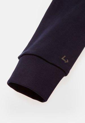 Granatowa koszula z długim rękawem z mankietem z materiału w prążki, z małym, beżowym detalem logo umieszczonym na dolnym brzegu. Gładka tekstura, dopasowany krój.