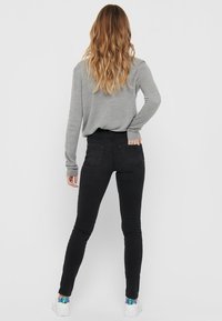 Schwarze Skinny-Jeans mit hoher Taille und zwei Gesäßtaschen, kombiniert mit einem grauen Langarmshirt. Das Modell trägt weiße Turnschuhe mit farbigen Akzenten.