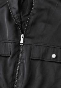 Veste noire avec fermeture éclair à l'avant et deux poches poitrine. Comprend un tissu lisse, du matériel en métal argenté et des éléments de design minimalistes.
