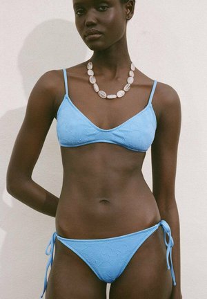 Light blue bikini set featuring a textured fabric, triangular top, and tie-side bottom. Accompanied by a chunky necklace.