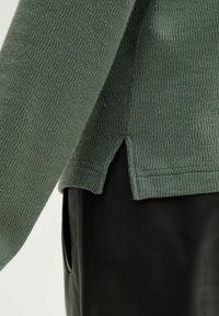 Groene gebreide trui met een geribbelde stof, voorzien van een zijsplit aan de onderkant, gedragen met een zwarte, gladde stof eronder.