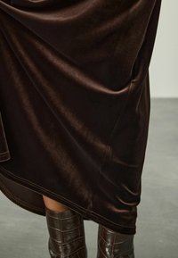 Gonna di velluto marrone scuro che si adagia sulle gambe abbinata a stivali di pelle a altezza ginocchio marrone scuro con motivo cocco su un pavimento grigio.