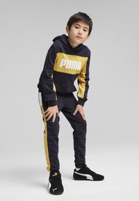 Tamsiai mėlyna džemperio ir kelnių komplektas su geltonais akcentais. Turtingas "PUMA" logotipas, raukšlėtos rankovės ir šoninės juostelės. Juodi sportbačiai su auksiniais akcentais.