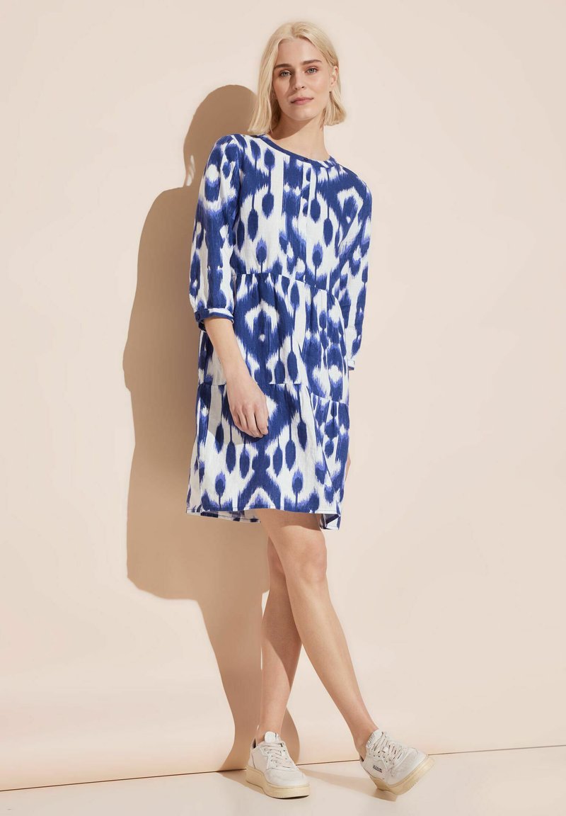 Blau-weißer, gemusterter Kleid mit Dreiviertelärmel, Taillenkordel, seitlichen Taschen und leichtem Stoff, getragen mit weißen Sneakers.