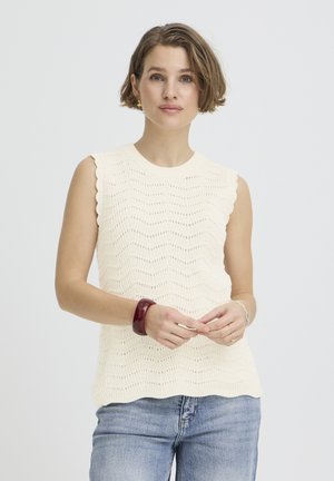 Kobieta o krótkich brązowych włosach, ubrana w kremowy top na ramiączkach z dzianiny, jasnoniebieskie dżinsy, złote kolczyki, czerwoną bransoletkę oraz srebrne pierścionki.