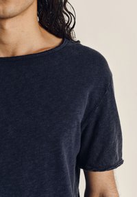 Person som bär en mörkblå t-shirt med korta ärmar, rund halsringning och råa sömmar, med långt mörkt lockigt hår synligt på ena sidan.