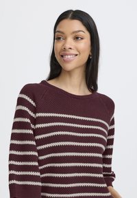 Knit sweater in burgundy with beige horizontal stripes. Features a round neckline and three-quarter raglan sleeves, textured fabric.