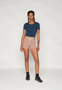 Woman with braided hair wearing a navy crop top, beige Nike Pro shorts, black socks, and black athletic shoes standing against a plain background.