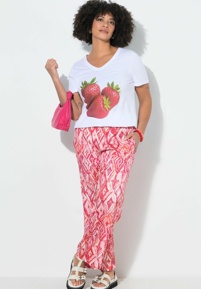 Frau trägt weißes T-Shirt mit großem Erdbeermotiv, rote gemusterte Hose, weiße Sandalen, hält rosa Handtasche und trägt ein rotes Armband.