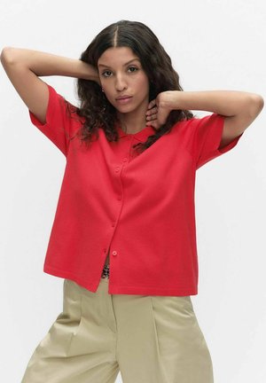 Vrouw met krullend haar, gekleed in een rood overhemd met korte mouwen en beige broek, handen achter haar hoofd geheven, neutrale uitdrukking.