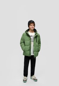 Groene gewatteerde jas met capuchon, ritssluiting aan de voorkant en drukknoopjes; gedragen over een witte graphic t-shirt en zwarte cargobroek; zwarte beanie.