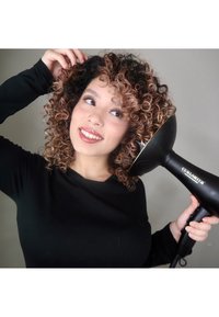 Czarna suszarka do włosów ze złotymi akcentami, duży okrągły dyfuzor, trzymana przez osobę z kręconymi włosami, ubraną w czarną bluzkę z długim rękawem.