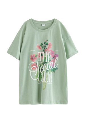 Ung kvinde iført en burgunder fleecetrøje, grøn t-shirt med grafik, mørke flared jeans og burgunder sko, stående mod en ensfarvet baggrund.