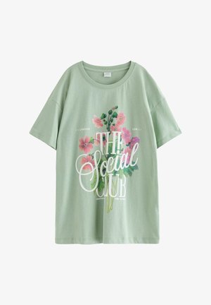Ung kvinde iført en burgunder fleecetrøje, grøn t-shirt med grafik, mørke flared jeans og burgunder sko, stående mod en ensfarvet baggrund.