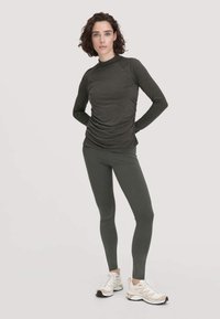 Dunkelgrünes Langarmoberteil mit Mock Neck, das mit Raffung an der Taille versehen ist. Kombiniert mit passenden dunkelgrünen Leggings und neutralen Sneakers.
