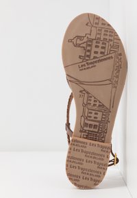 Sandale marron avec une tige en cuir texturé, une sangle ajustable et une semelle imprimée présentant des motifs de paysage urbain et la marque "Les Tropéziennes."