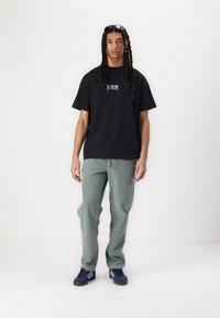Tricou din bumbac negru cu grafică pe față, asortat cu pantaloni verzi deschis. Modelul poartă adidași albaștri și ochelari de soare. Design simplu, croială lejeră.