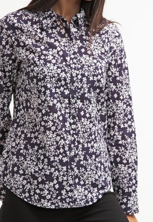 Femme portant une chemise bleu marine à boutons avec un motif floral blanc et des manches longues, assortie à un pantalon noir.