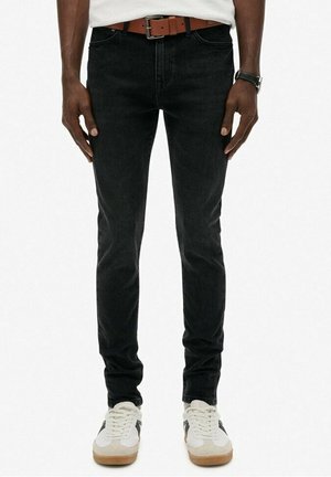 Mężczyzna noszący czarne jeansy typu slim fit, brązowy pasek, białe tenisówki z czarnymi paskami i białą koszulę z zegarkiem na lewej ręce.